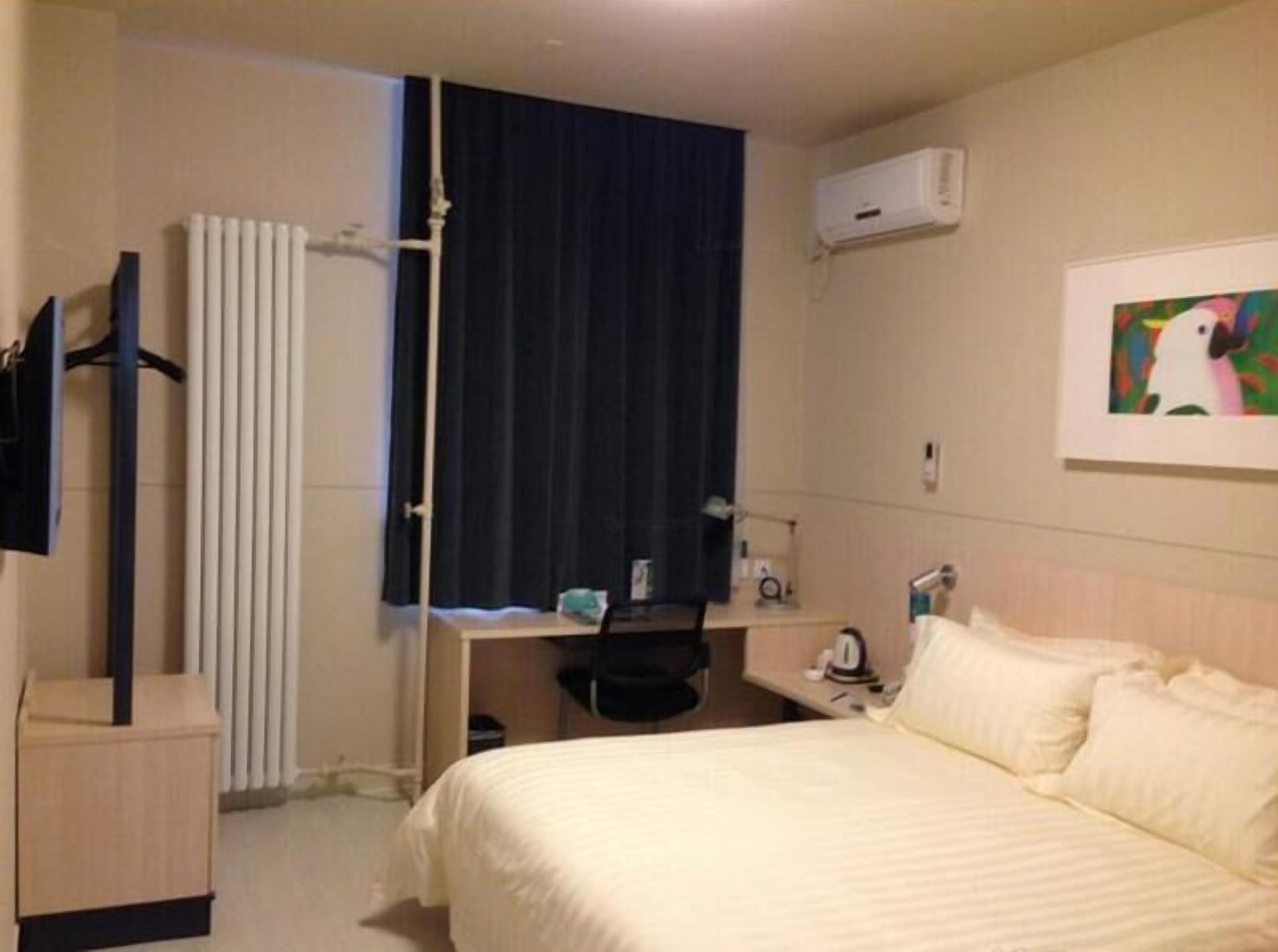 A hotel room with a bed, a desk, a chair, a radiator, an air conditioner, and a painting on the wall.