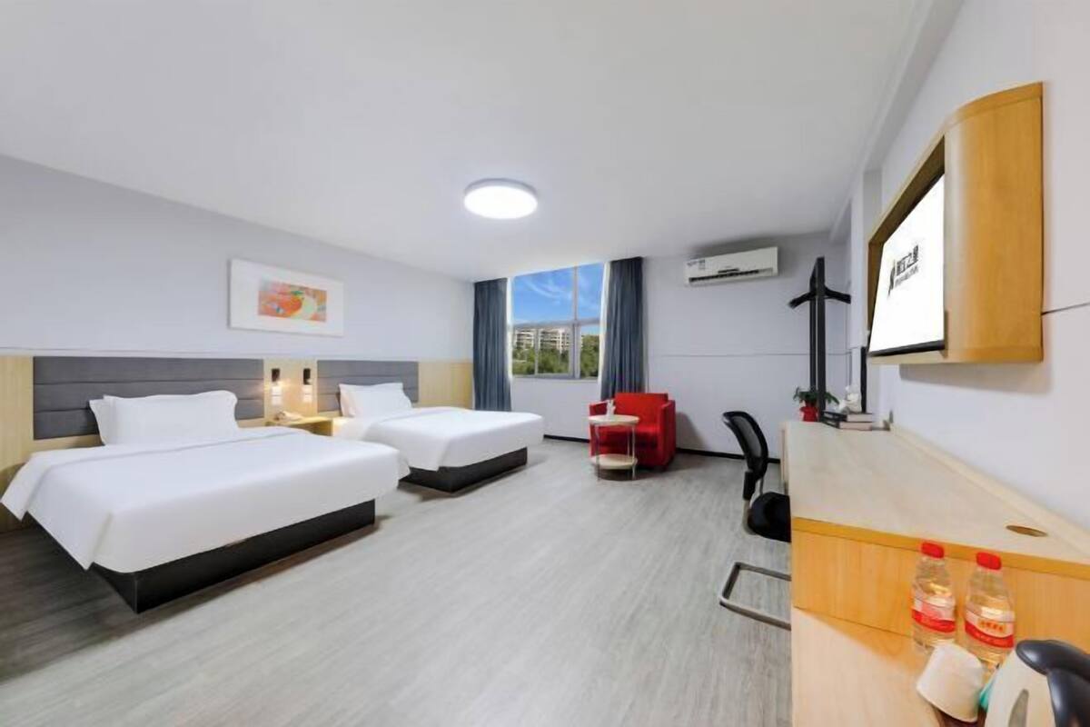 Une chambre d’hôtel avec deux lits, un bureau avec une chaise, une chaise rouge, une télévision et une fenêtre donnant sur des immeubles.