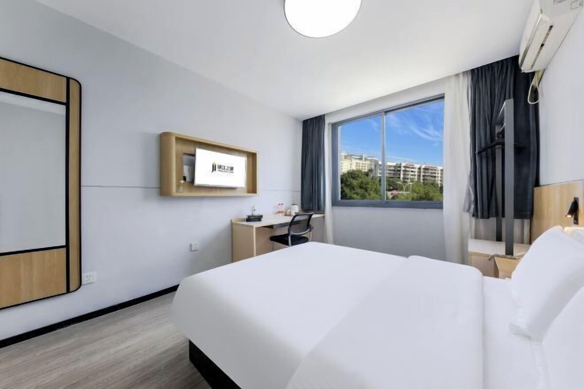 A hotel room with a large bed, a desk, a chair, a mirror, and a window with a city view.