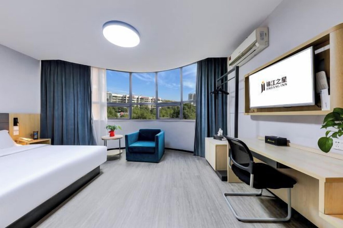 A hotel room with a bed, a desk, a chair, a small table, a potted plant, and a large window with a view.