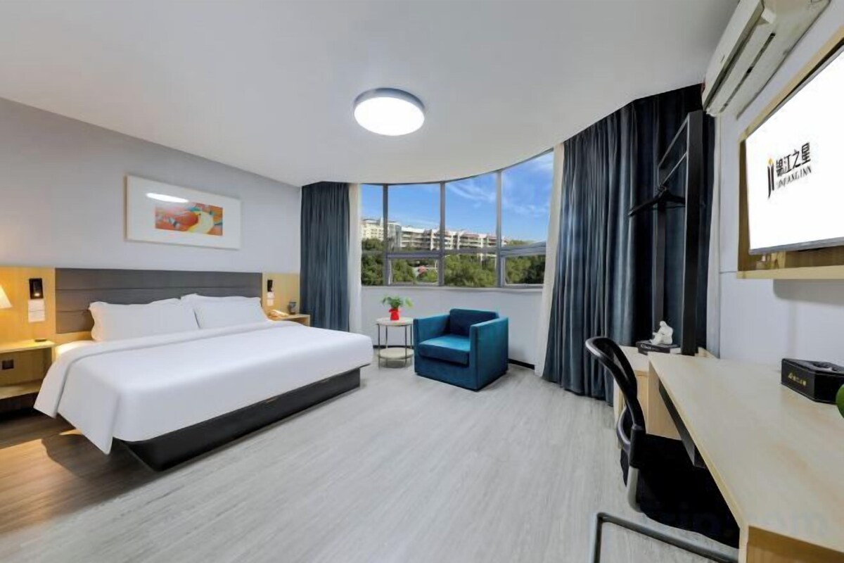 Ein Hotelzimmer mit einem großen Bett, einem Schreibtisch, einem Sessel, einem kleinen Tisch und einem Fenster mit Vorhängen.