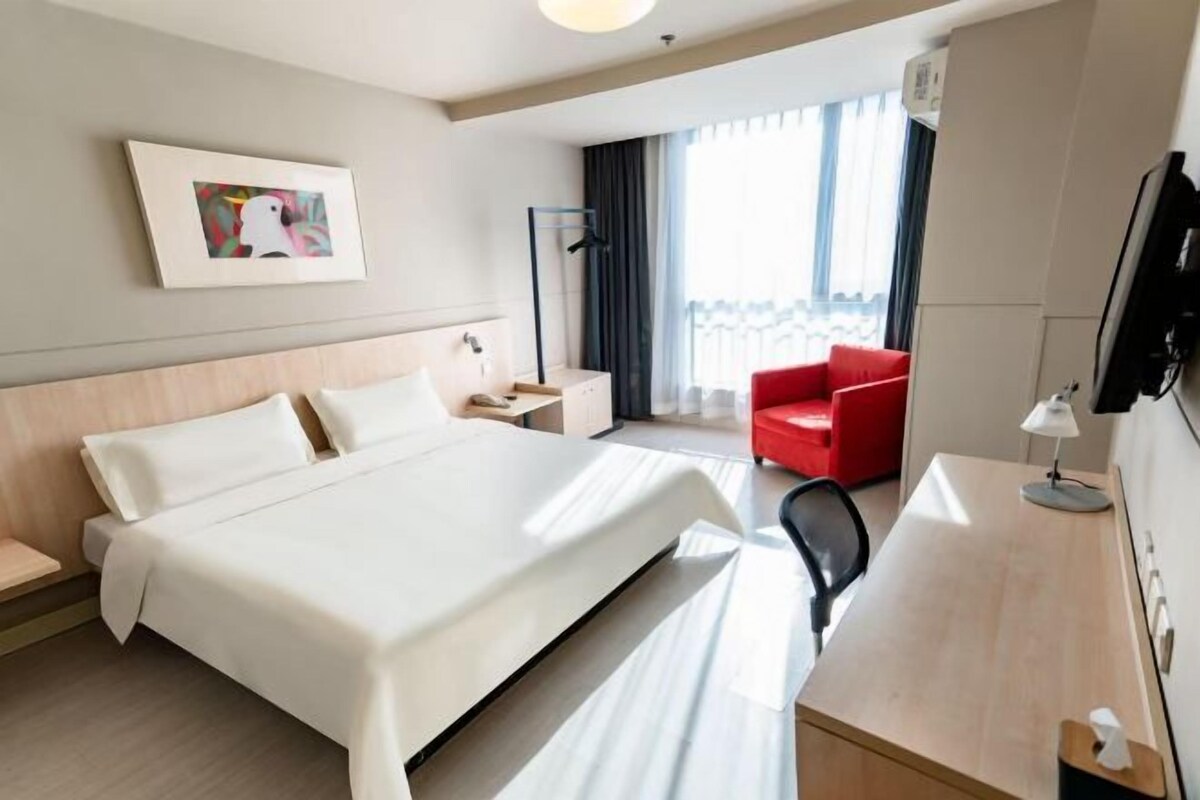 A hotel room with a bed, a desk, a chair, a red armchair, a TV, and a window with curtains.