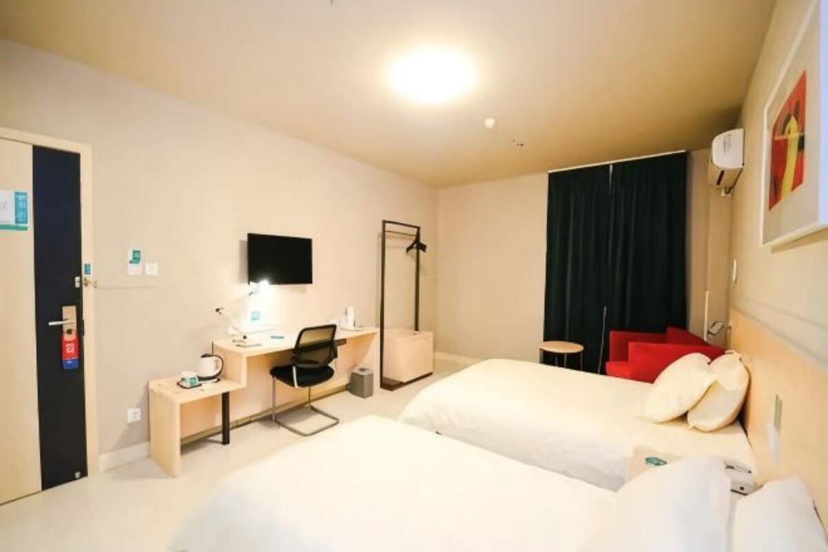 A hotel room with two beds, a desk with a chair, a desk lamp, a flat-screen TV, a mirror, and a red chair.