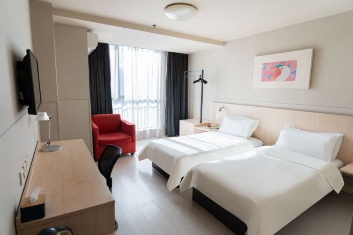 A hotel room with two beds, a desk, a chair, a TV, a bedside table, a lamp, and a window with curtains.