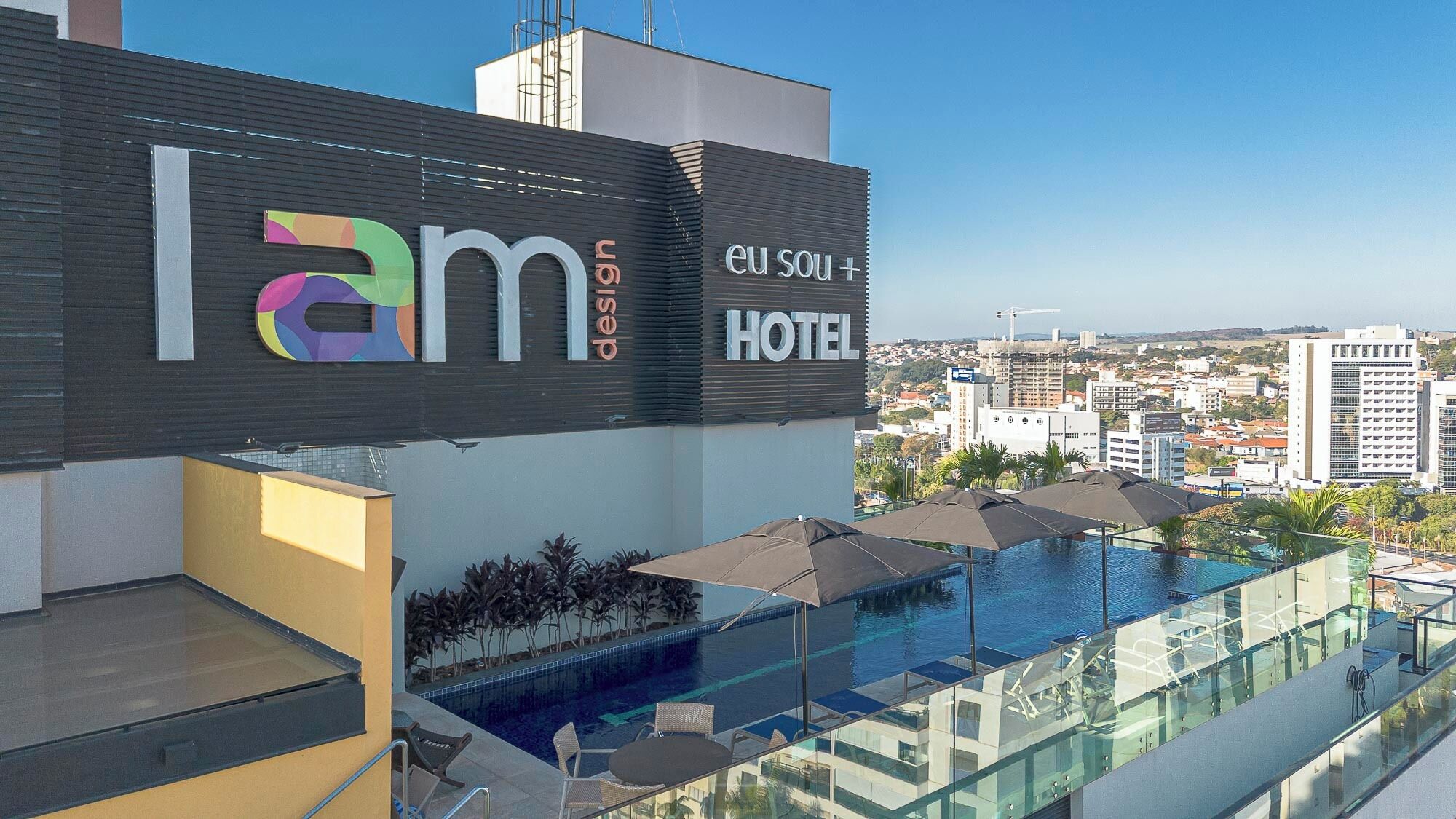 Letrero de hotel con la marca 'lam' y 'eu sou +', piscina en la azotea con sillas de descanso y vistas a la ciudad.