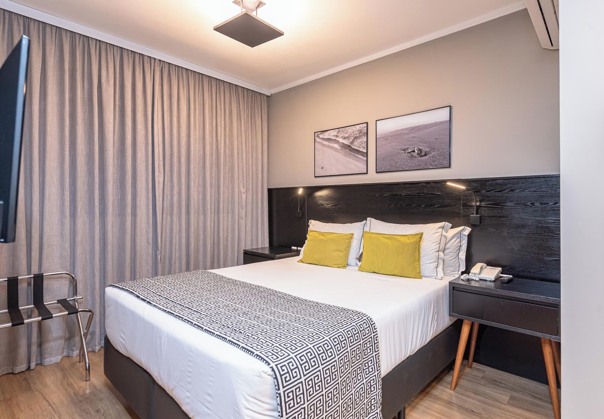 Une chambre d’hôtel moderne avec un grand lit, une table de chevet avec une lampe, une chaise et deux tableaux encadrés au mur.