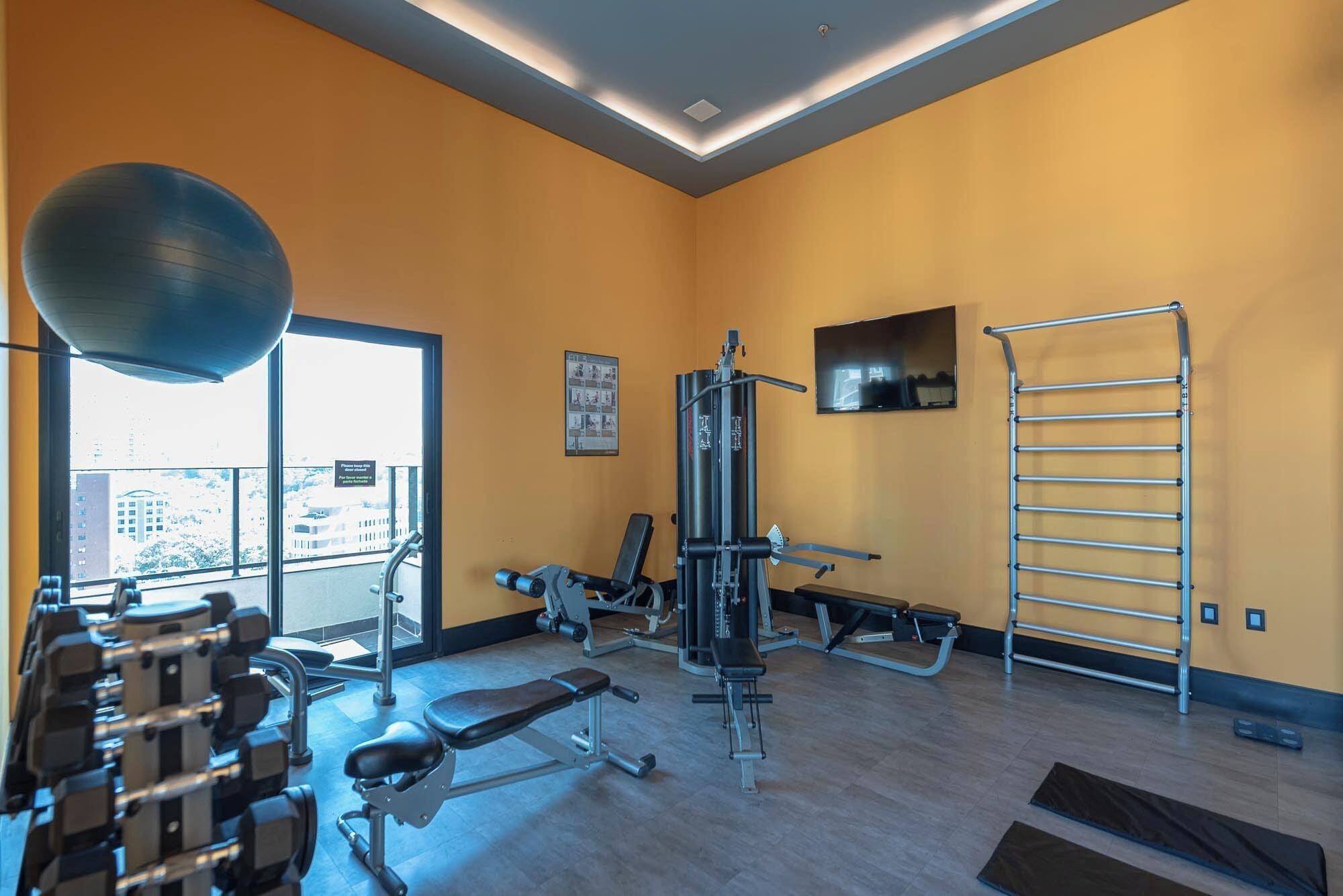 A gym with exercise equipment, a bench, a wall-mounted TV, and a window with a view of buildings.