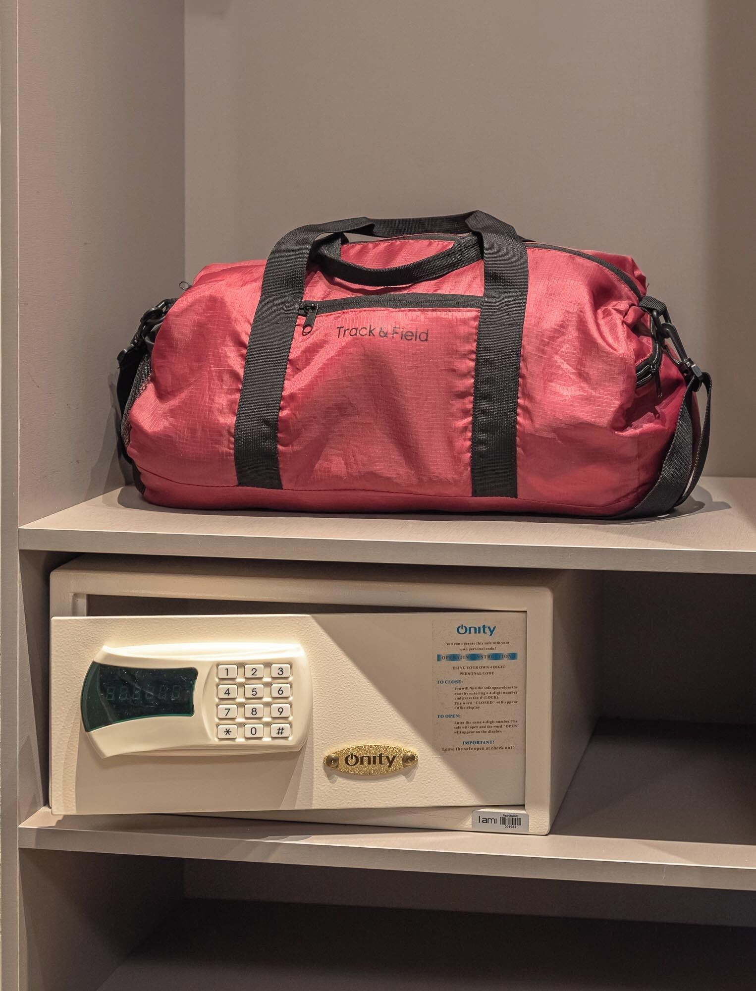 A red duffel bag labeled "Track & Field" on a shelf above a white security keypad with a gold "Unity" keypad and a sign with instructions.