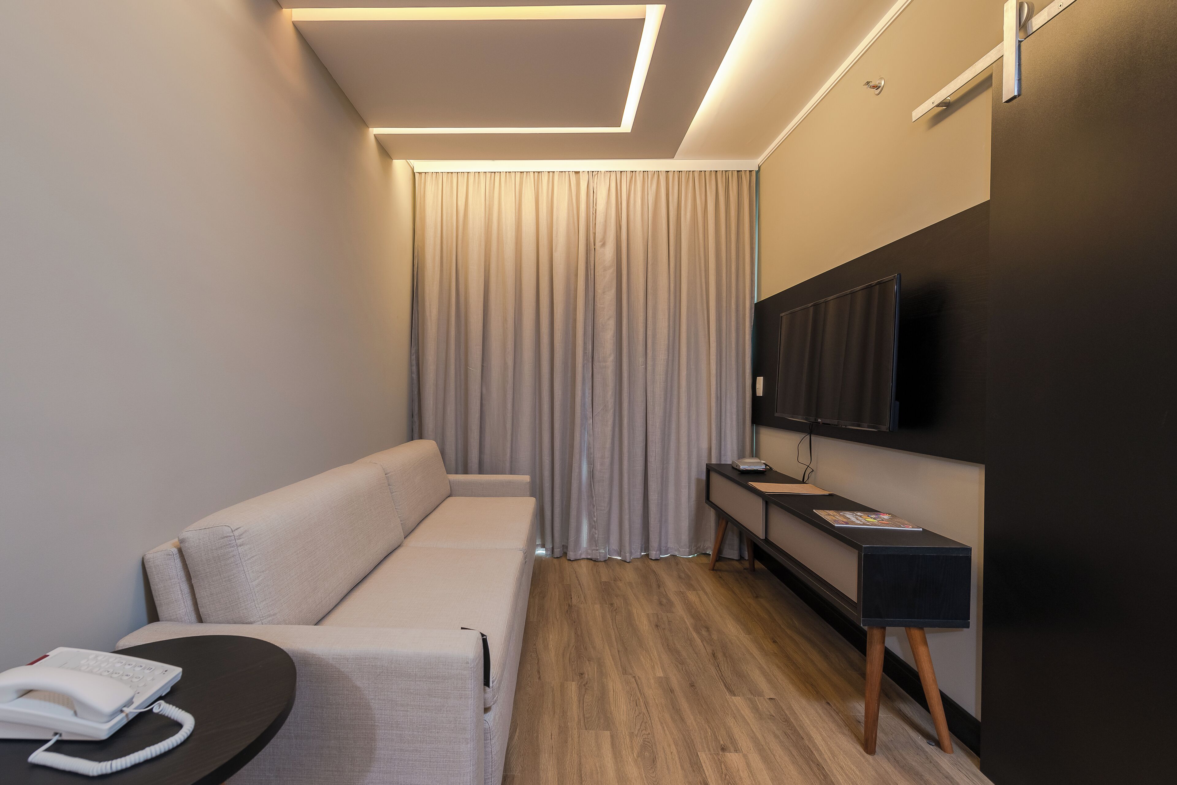 Habitación de hotel moderna con sofá, una mesita con teléfono, televisor en la pared y piso de madera.