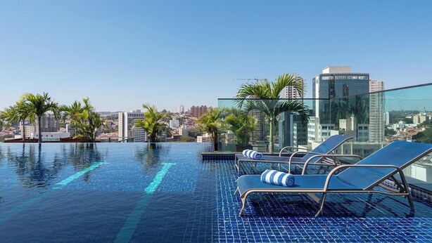 Una piscina infinita en la azotea con sillones de descanso y palmeras, con vista a un paisaje urbano con edificios modernos.