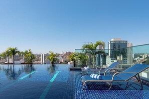 Una piscina infinita en la azotea con sillones de descanso y palmeras, con vista a un paisaje urbano con edificios modernos.