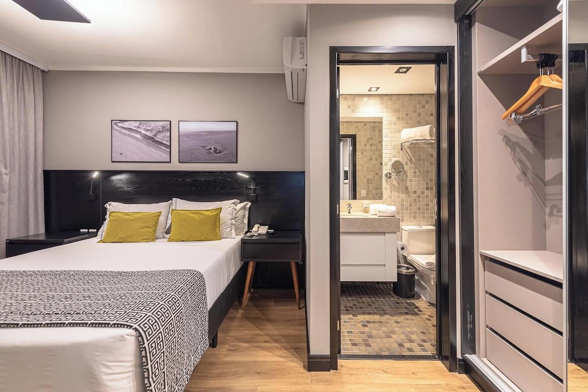 Habitación de hotel moderna con una cama grande, mesitas de noche, un armario y un baño visible a través de una puerta abierta.