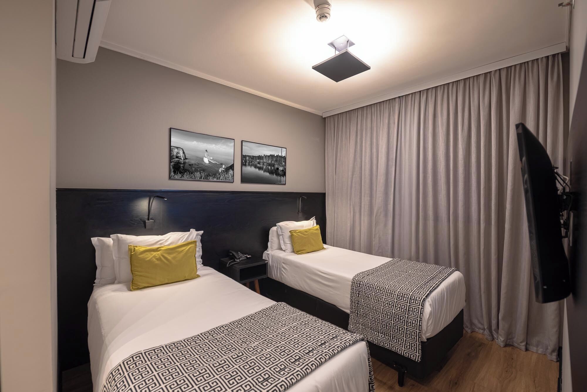Habitació d'hotel amb dos llits, un capçal fosc, una làmpada al sostre i dos quadres al mur.