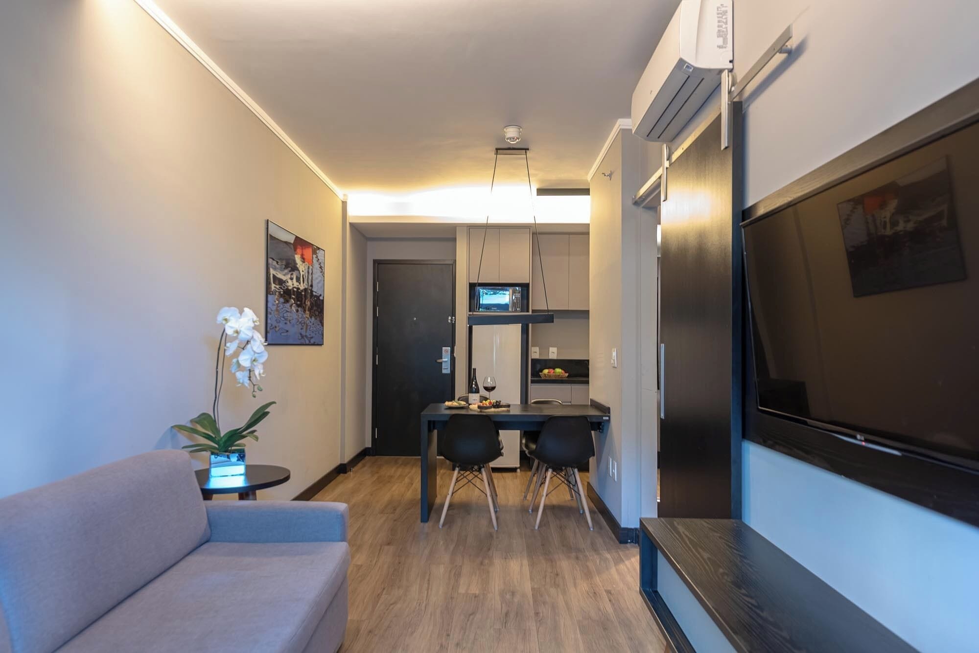 A compact living space with a sofa, a dining table with chairs, a small kitchen area with a microwave, and a TV mounted on the wall.