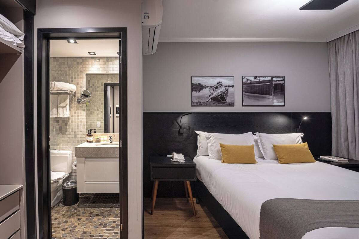 Una habitación de hotel moderna con una cama grande, una mesita de noche, un tocador, un baño con lavamanos y espejo, y dos cuadros en la pared.