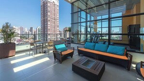 Terraza en la azotea con mobiliario exterior, un muro de cristal y vistas a un edificio alto.