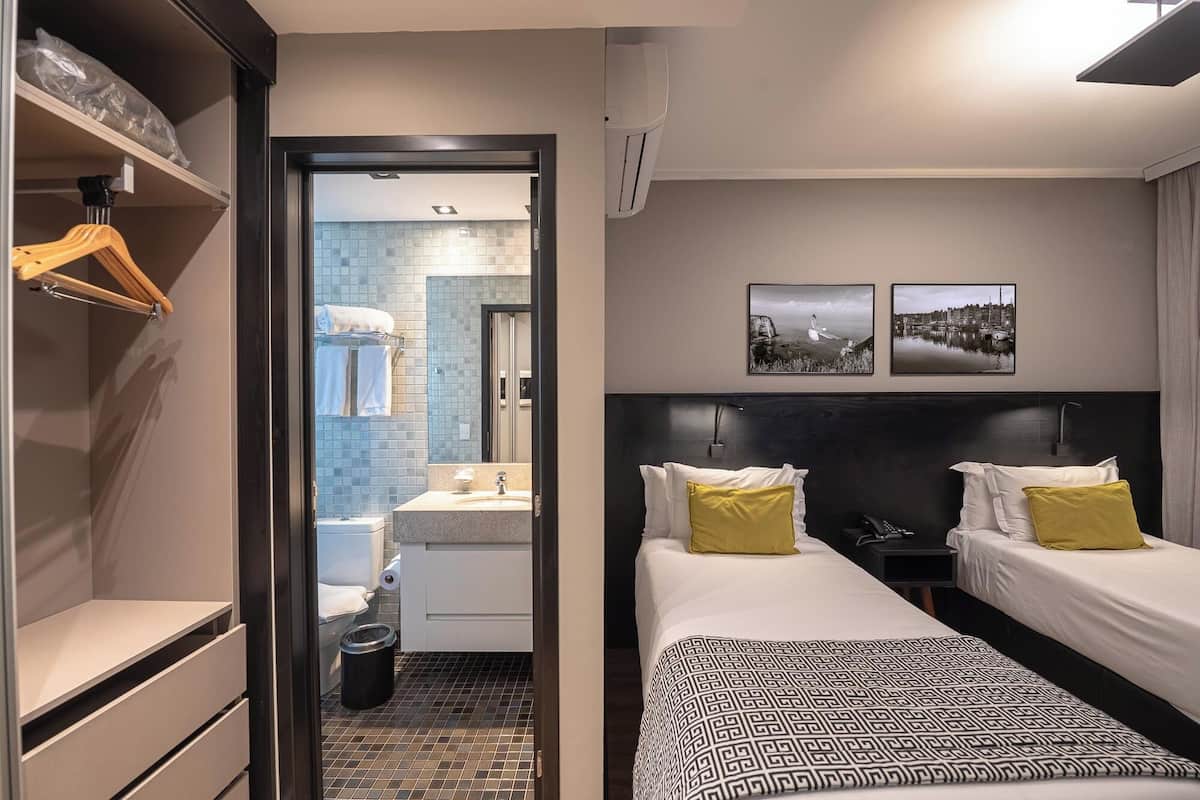 Habitación de hotel con cama, mesitas de noche, armario, baño con lavabo e inodoro, y dos cuadros colgados en la pared.