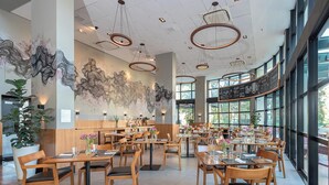 Un restaurante espacioso con mesas y sillas de madera, ventanas amplias e iluminación colgante moderna.