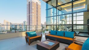 Una terraza en la azotea con mobiliario de mimbre, un muro de cristal y vistas a un edificio alto.