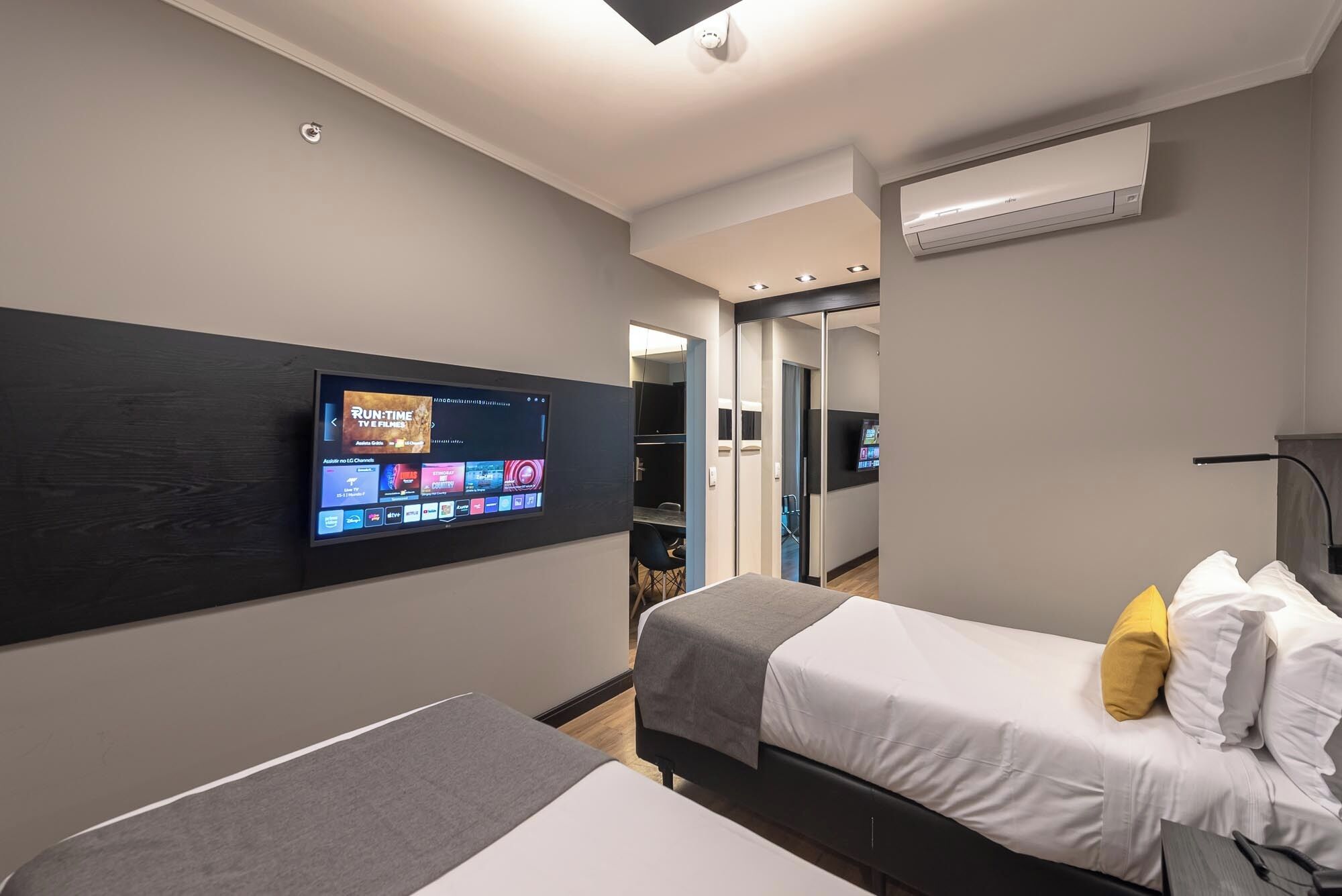 Habitación de hotel con televisión de pantalla plana montada en la pared, dos camas, una mesita de noche y un equipo de aire acondicionado.