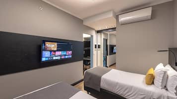 Habitación de hotel con televisión de pantalla plana montada en la pared, dos camas, una mesita de noche y un equipo de aire acondicionado.
