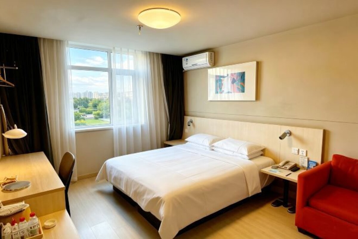 Habitación de hotel con cama, escritorio y silla, un sillón rojo, una ventana con cortinas y un equipo de aire acondicionado.
