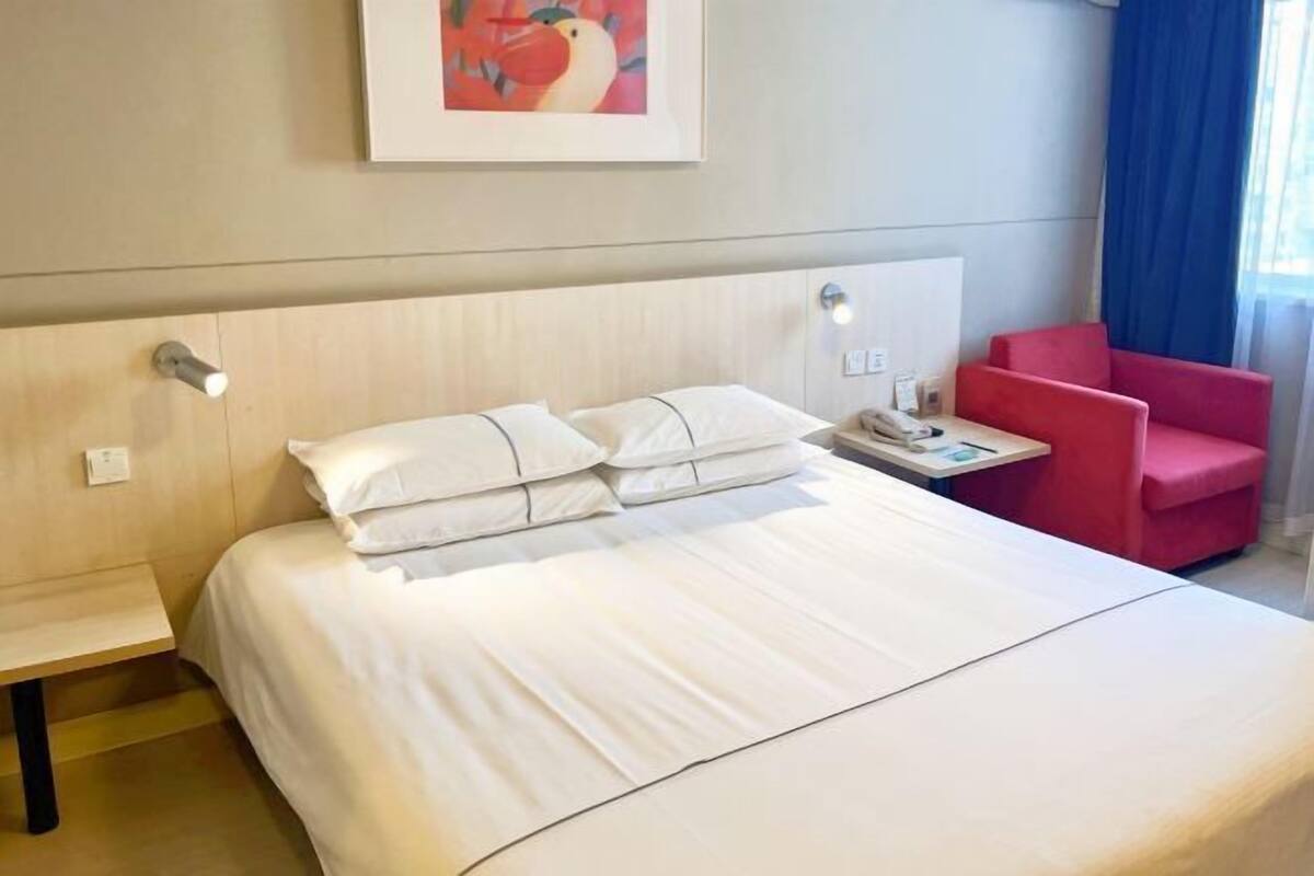 Hotellihuoneessa on sänky, yöpöytä lampun kanssa, punainen tuoli ja seinällä oleva kehystetty kuva.