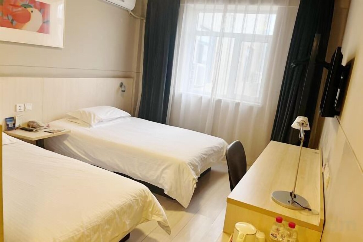 A hotel room with two beds, a bedside table with a lamp, a small desk, and a window with curtains.