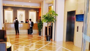 Een hotelhal met een marmeren vloer, een receptiebalie, liften en potplanten.