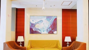 Een hal met een gele bank, twee bruine fauteuils, een ronde salontafel, en een groot abstract schilderij aan de muur.