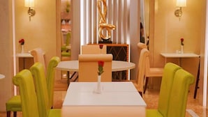 Een restaurantgedeelte van een hotel, met een ronde tafel, vier groene stoelen en een witte tafelkleed. Er zijn twee wandlampen, een decoratieve gouden sculptuur en een vaas met een rode roos op de tafel.