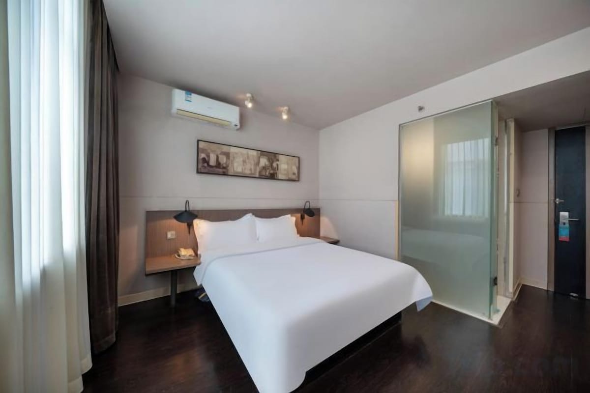 A hotel room with a large bed, a bedside table, a nightstand, a wall-mounted air conditioner, and a sliding glass door.
