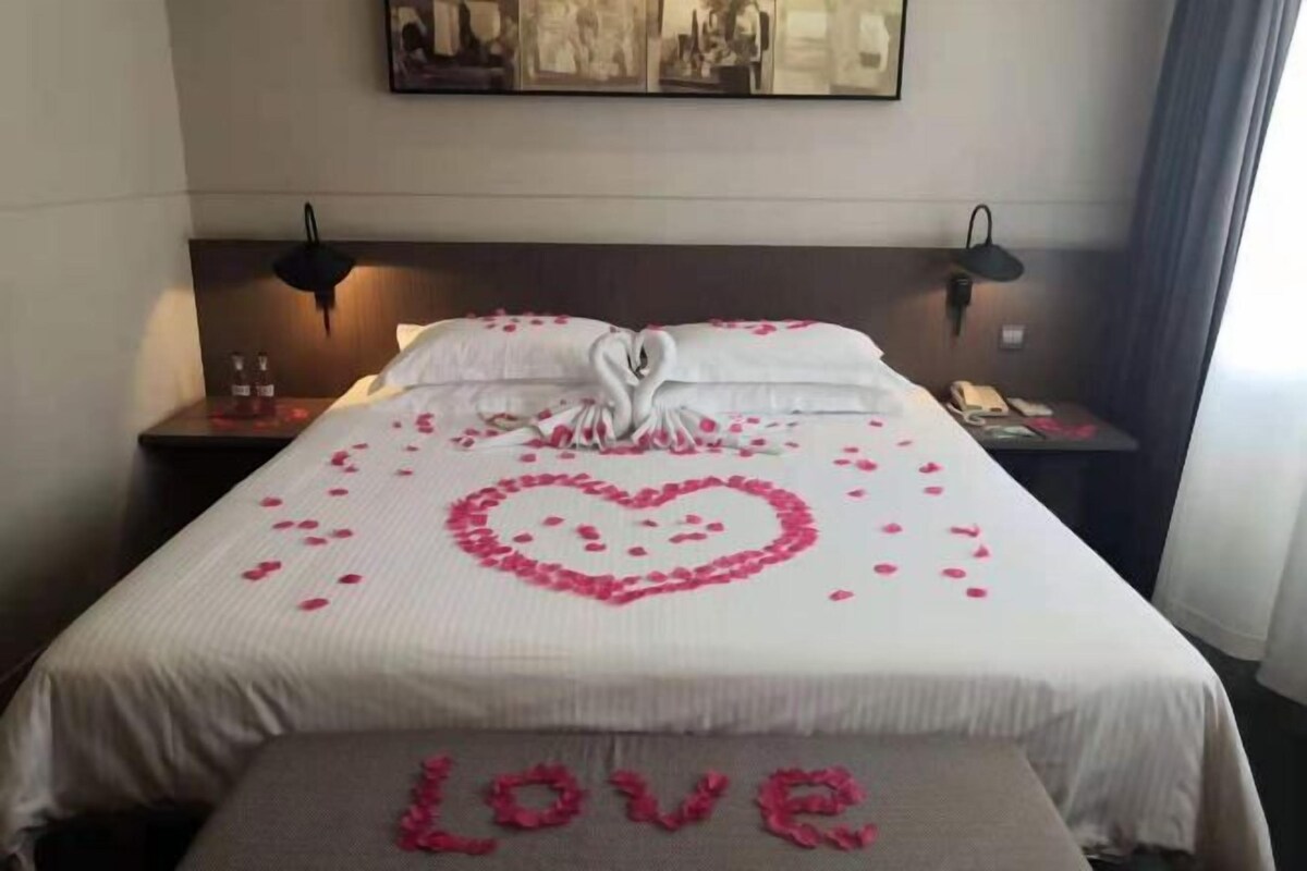 Une chambre d’hôtel avec un lit parfaitement fait, une décoration en forme de cœur posée sur le lit, et un banc sur lequel est inscrit le mot « AMOUR » à l’aide de pétales de rose.