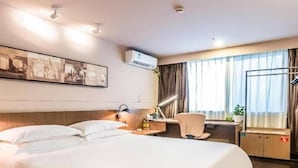 A hotel room with a large bed, a desk with a chair, an air conditioning unit, and a window with curtains.