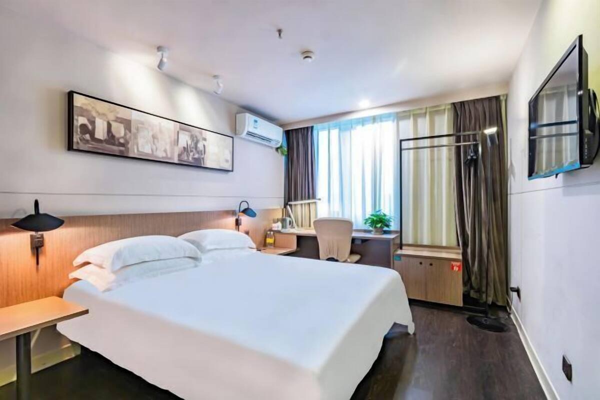 A hotel room with a large bed, a desk with a chair, a TV mounted on the wall, and a window with curtains.
