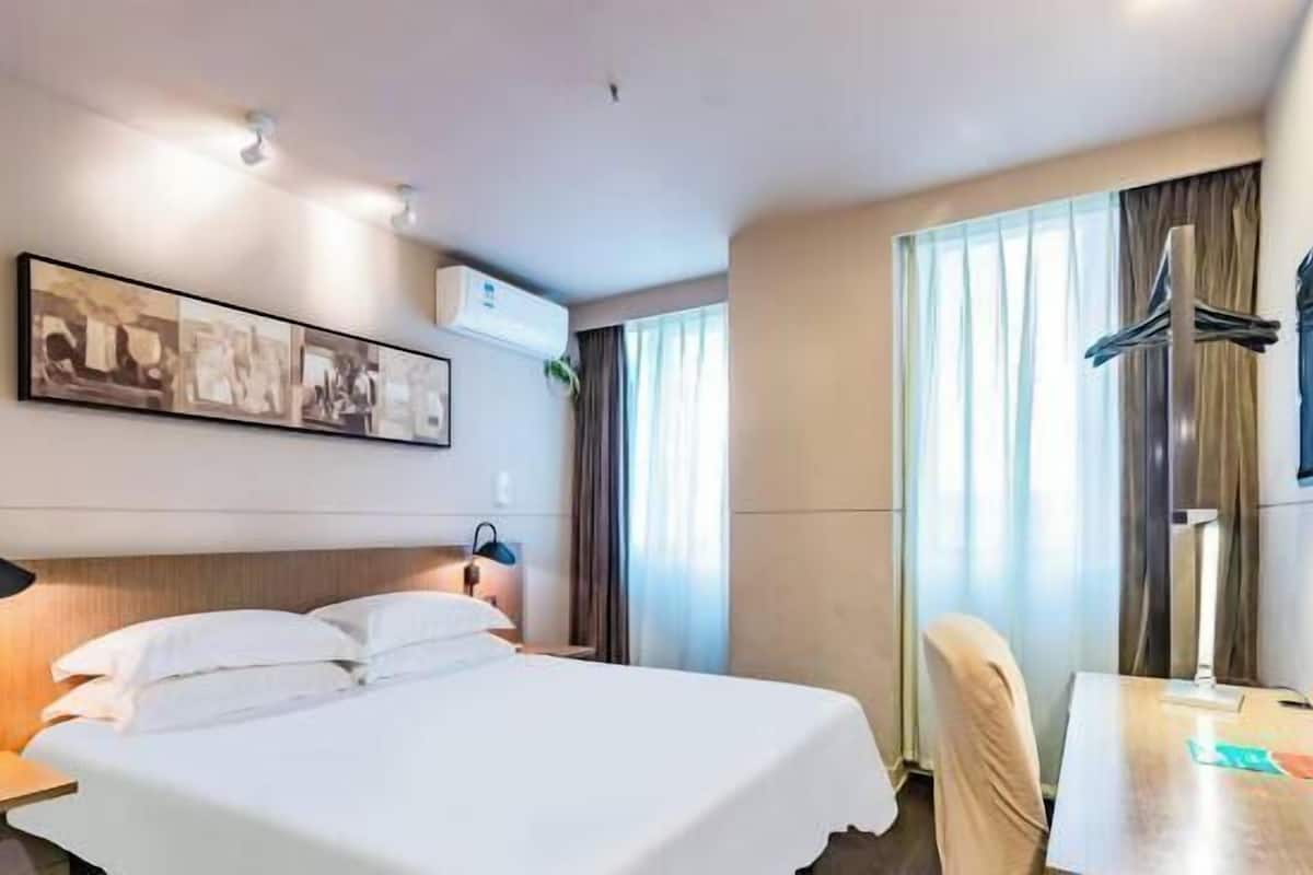 A hotel room with a bed, a desk, a chair, a lamp, a coat rack, and a wall-mounted air conditioner.