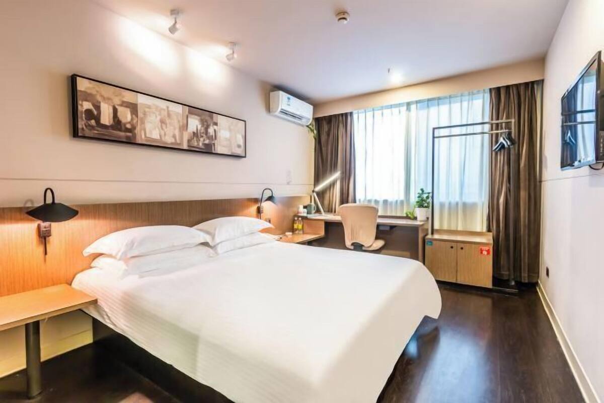 A hotel room with a bed, bedside table, lamp, chair, desk, and air conditioning unit.