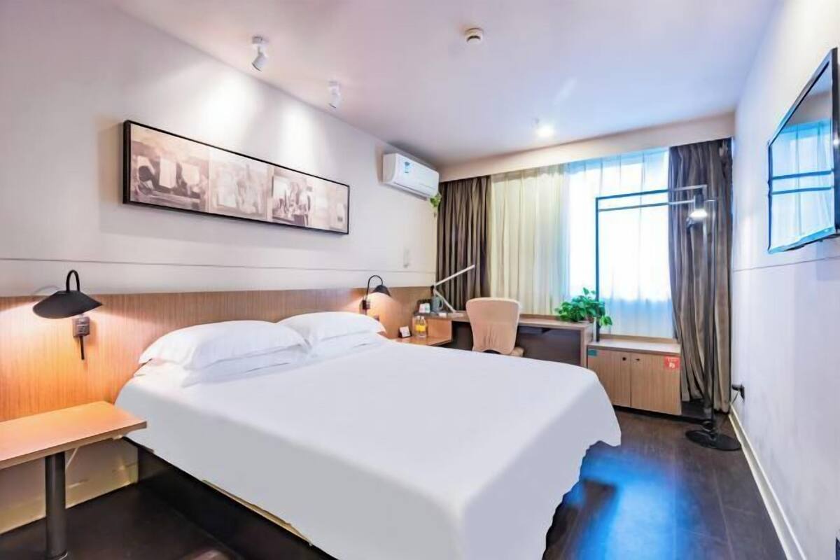 A hotel room with a bed, bedside tables, a desk, a chair, a window with curtains, and a wall-mounted TV.