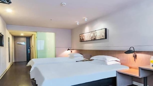 A hotel room with two beds, a bedside table, a wall-mounted lamp, and a ceiling-mounted smoke detector.