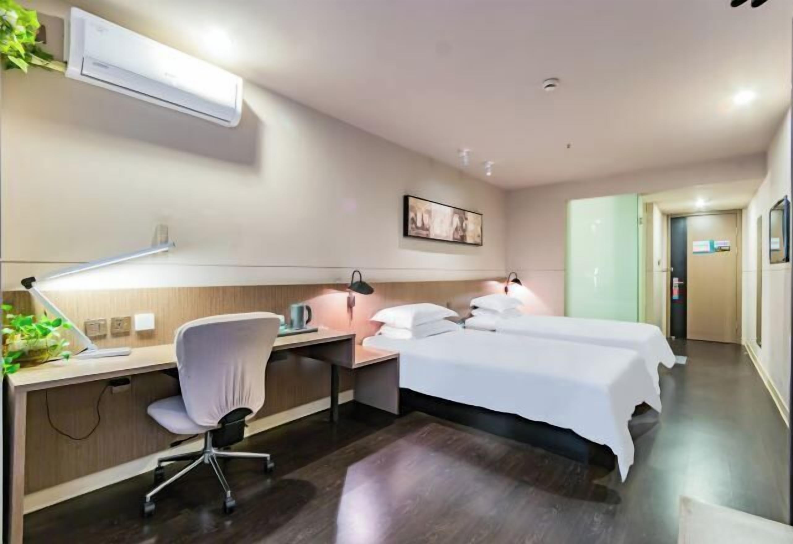 Habitación de hotel con escritorio, silla, dos camas, aire acondicionado de pared, un cuadro enmarcado y una puerta que da a otra habitación.