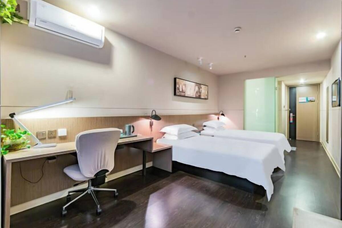 A hotel room with a desk, chair, two beds, a wall-mounted air conditioner, a framed picture, and a door leading to another room.