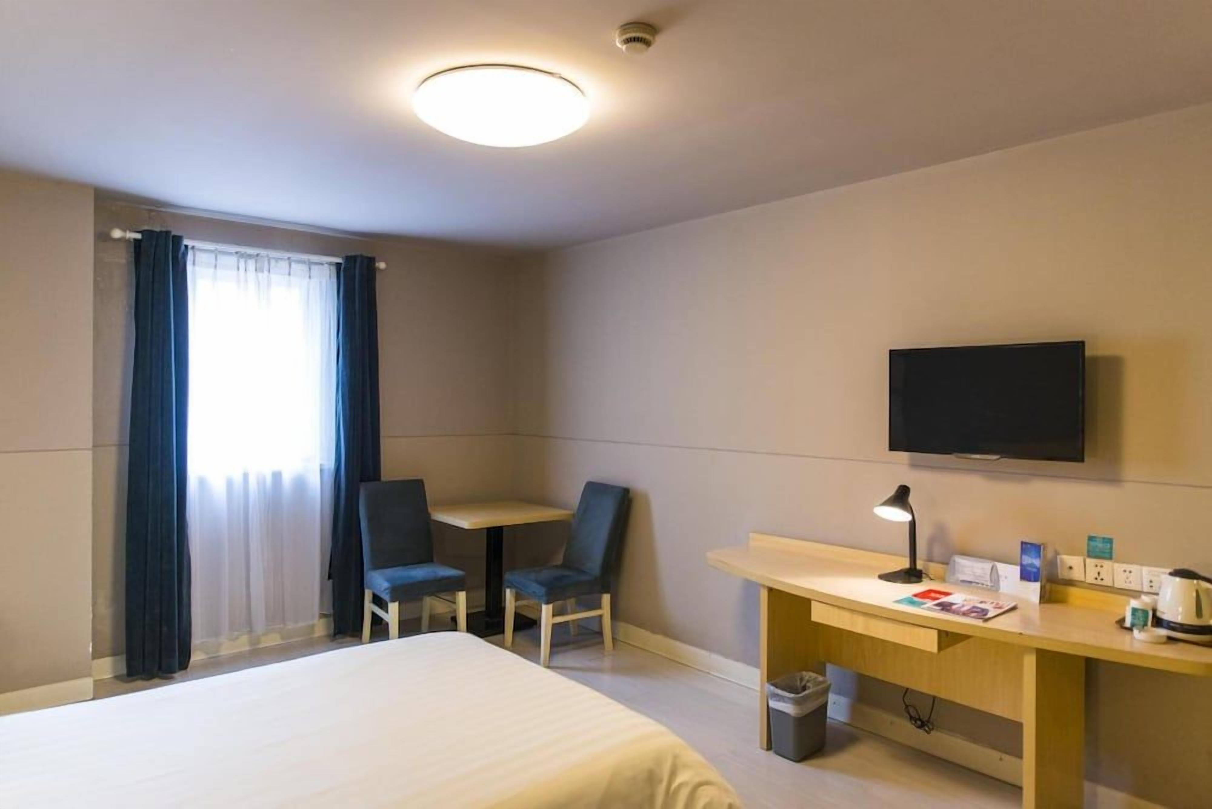A hotel room with a bed, a desk, two chairs, a television, and a window with curtains.