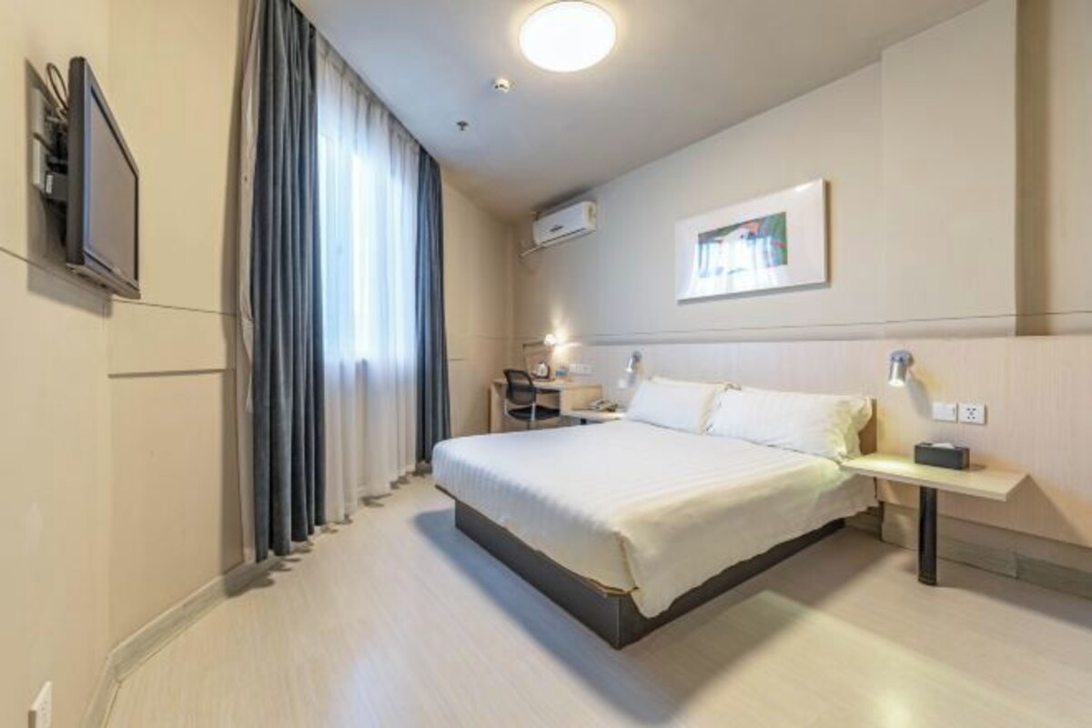 Una habitación de hotel con cama, mesita de noche, lámpara, aire acondicionado y televisor en la pared.