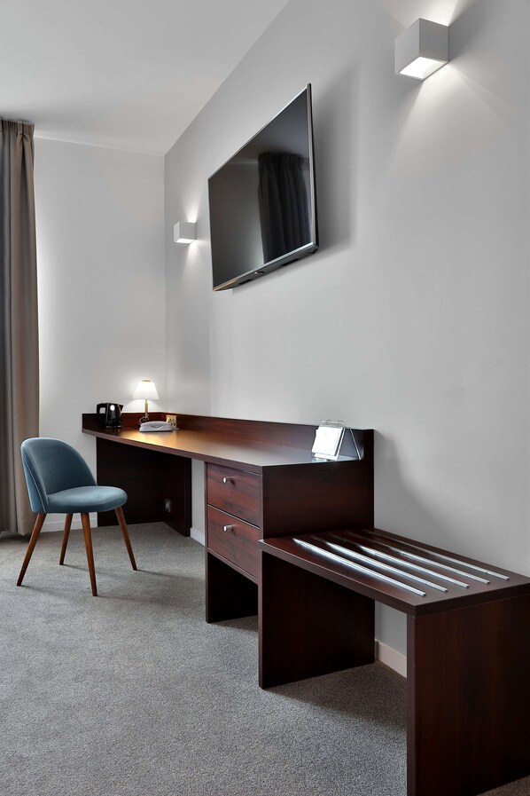 Una habitación de hotel moderna con un escritorio de madera oscura, una silla azul, un televisor de pantalla plana y una lámpara de escritorio.
