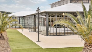 A covered outdoor area with a glass enclosure, palm trees, and a white building in the background.