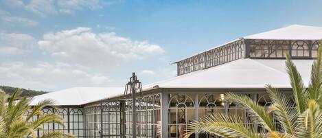 A covered outdoor area with a glass enclosure, palm trees, and a white building in the background.