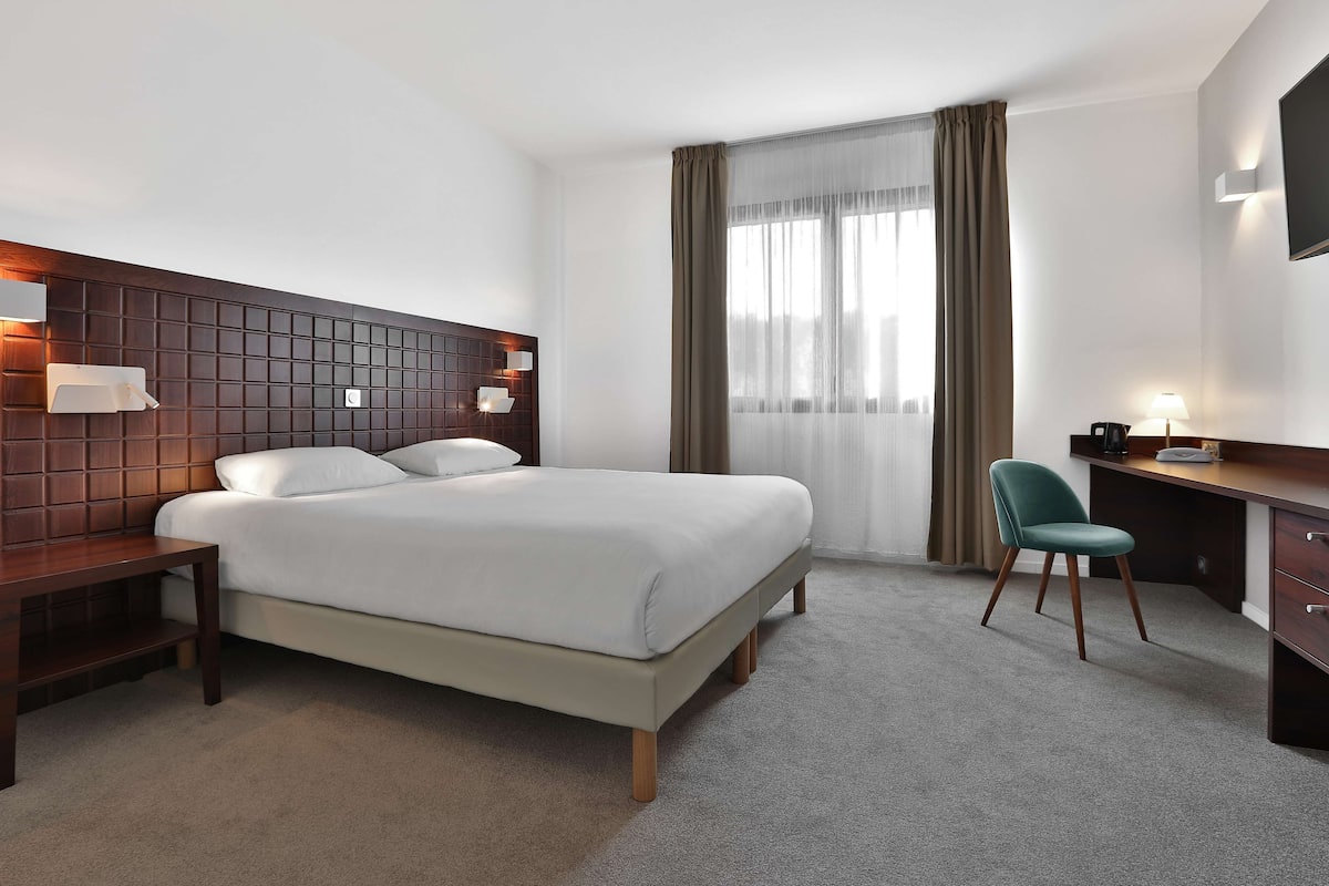 Ein modernes Hotelzimmer mit einem großen Bett, einem Schreibtisch, einem Sessel und einem an der Wand montierten Fernseher.