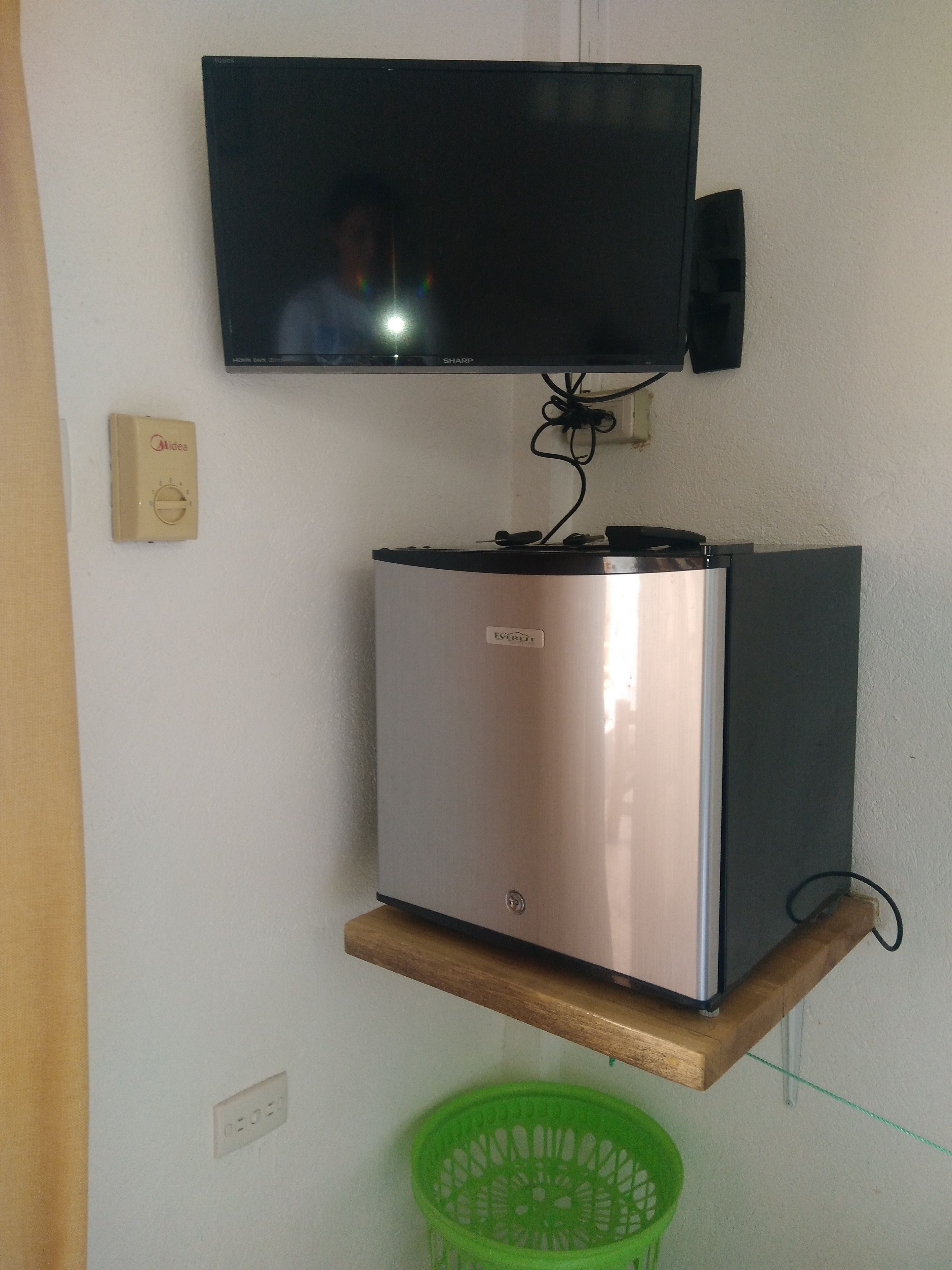 A compact refrigerator on a wooden shelf, a wall-mounted television, and a power outlet with a switch.