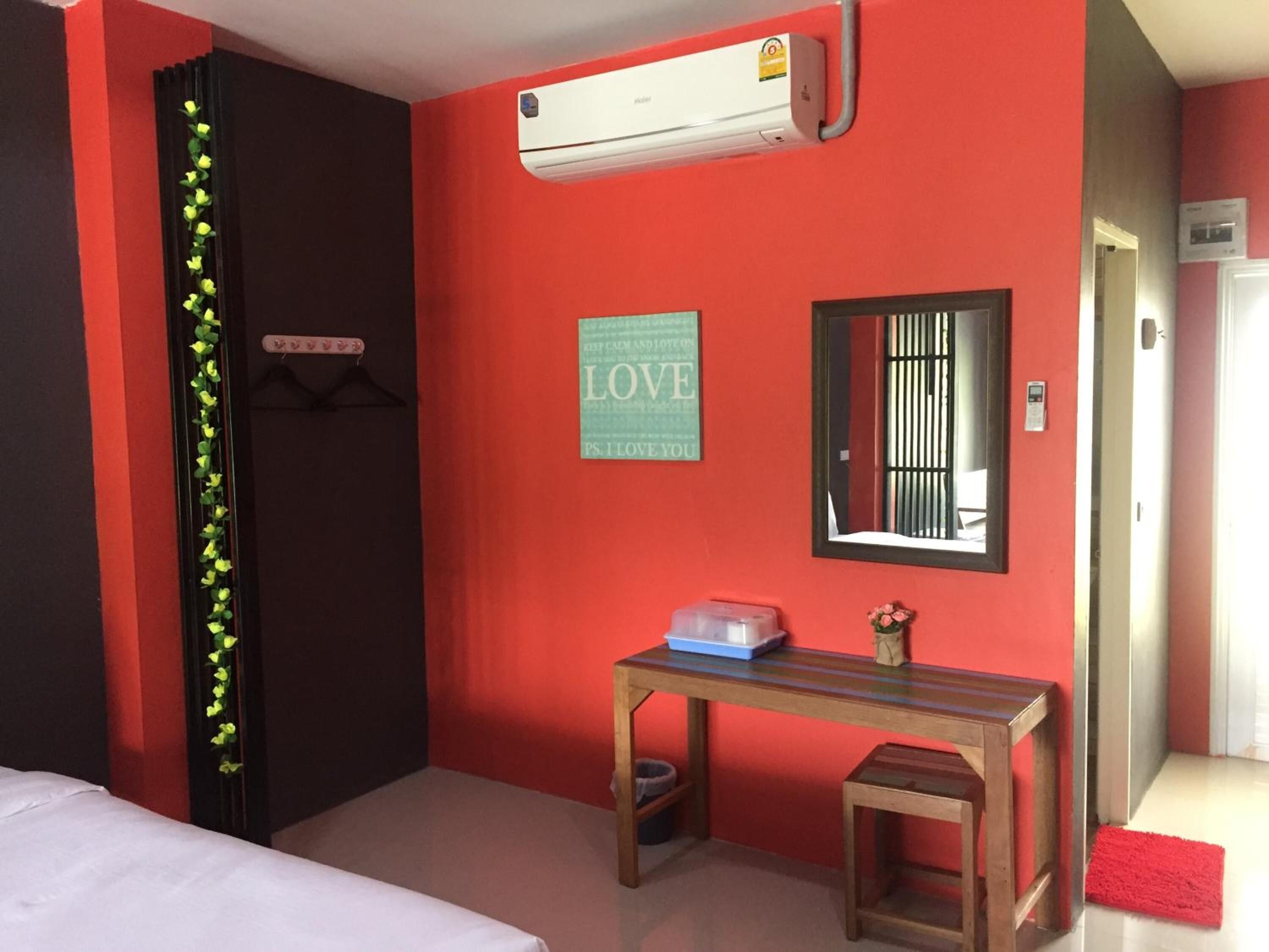 A hotel room with a red wall, a bed, a desk with a laptop, an air conditioner, a mirror, and a small table with a vase.