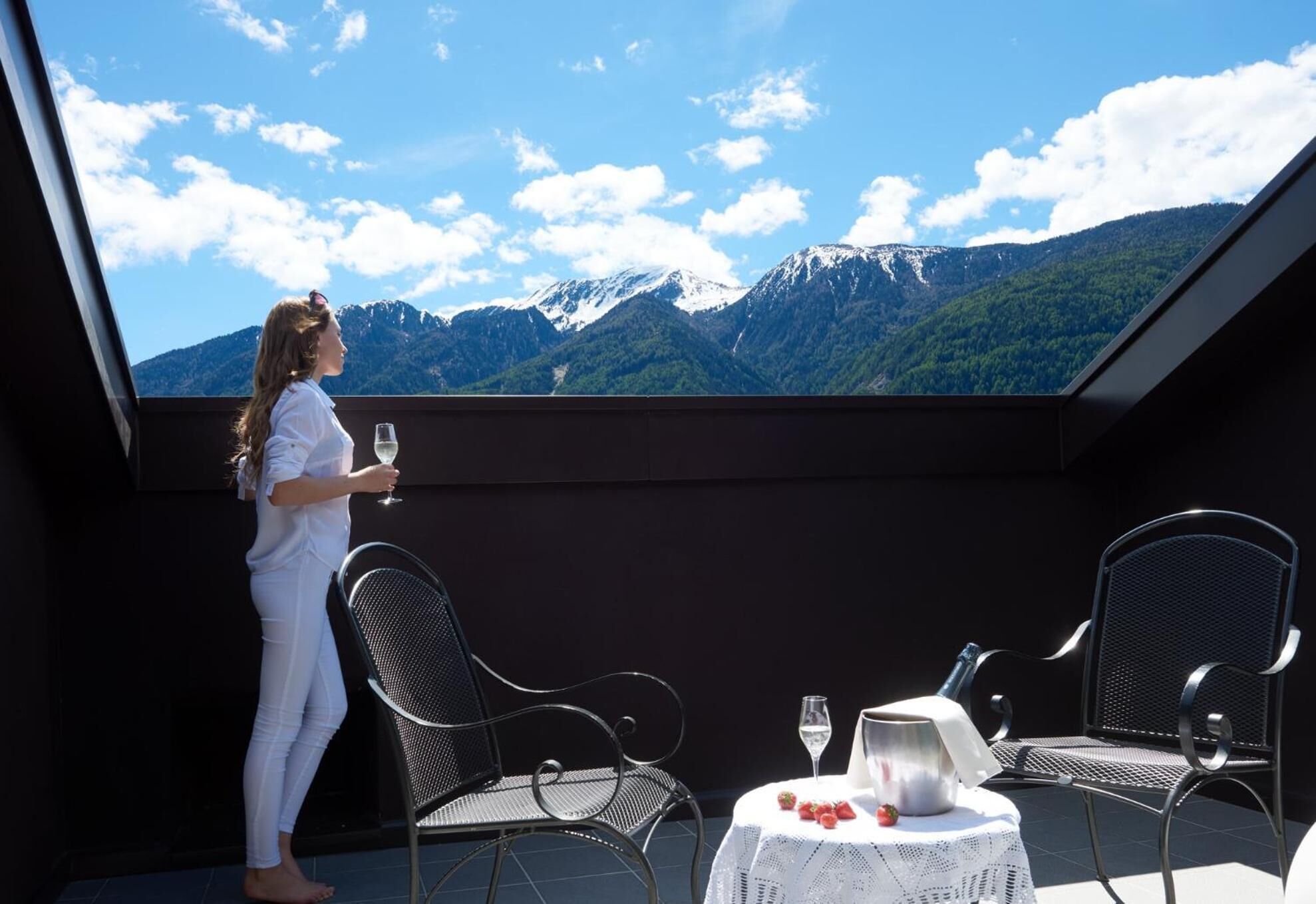 Ein Balkon mit Bergblick, zwei Stühle, ein Tisch mit einer Flasche und Gläsern sowie eine Person, die ein Glas hält.